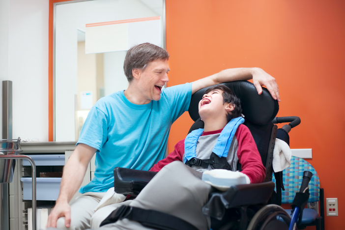 Young boy laughing while sitting in a wheelchair and father looking happily at him.